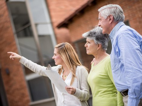 Immobilienmakler werden, Junge Immobilienmaklerin zeigt älterem Paar ein Gebäude von außen. Foto: iStock.com/andresr